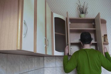 Genç adamın ahşap bir mutfakta önlüksüz görüntüsü. Kafasında ıslak tepsi var. Erkek yiyeceği boş karyola tahtasıyla arıyor. Kapıları aç. 