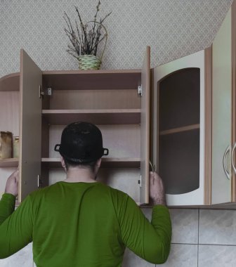 Genç adamın ahşap bir mutfakta önlüksüz görüntüsü. Kafasında ıslak tepsi var. Erkek yiyeceği boş karyola tahtasıyla arıyor. Kapıları aç. 