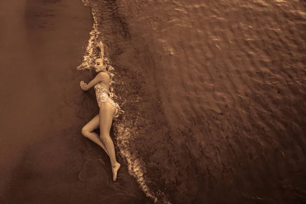 漂亮的性感女模特躺在白色沙滩上 穿着黑色泳衣 有着晒黑的身体 长长的金发 丰满的胸部 健康的大自然 一个年轻的女人穿着白色的泳衣躺在海边的沙滩上 — 图库照片
