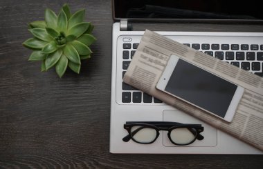 Laptoplu çalışma masası ve bir sürü masa aksesuarıyla çevrili mesaj için boş yer. Boş ekranlı cep telefonu, yeşil bitki, gözlük, gazete. Üst Manzara. Yazı için boş kopyalama alanı.