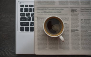 Bir fincan kahve, gazete ve dizüstü bilgisayarla iş masasının en üst görüntüsü. Bulanık metin. Yeni haberler. 
