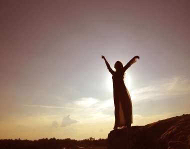 Mavi gökyüzü arka planına karşı bir dağın yamacında duran şehvetli seksi kadının silueti. Eller yukarı. gün batımı ışığı. 