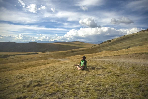 Young tourist  relaxing in the mountains, away from the city life and business meetings. Lonely man in green hoodie, sitting on the top of mountain hill under dramatic cloudy sky in high grass and enjoying of tranquility and solitude. Mountain Altai