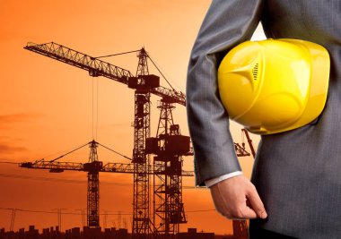 Yeni binanın arka planında işçiler için miğfer tutan mühendis ve akşam üstü bulutlu gökyüzünün arka planında inşaat vinçleri.