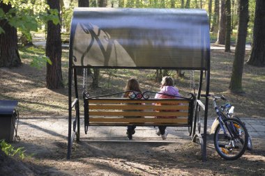Simple life without technology - two school girls wearing jacket  are riding on a swing in a playground before afternoon classes begins with happiness. back, rear view. bicycle stand near.