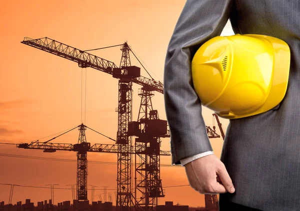engineer holding  helmet for workers security on background of new  building and construction cranes on background of evening sunset cloudy sky
