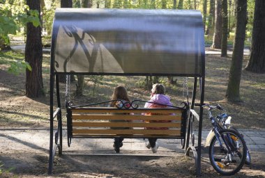 Simple life without technology - two school girls wearing jacket  are riding on a swing in a playground before afternoon classes begins with happiness. back, rear view. bicycle stand near.