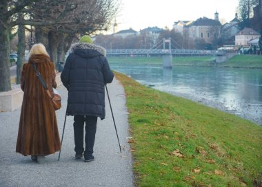 Yaşlı bir çift kışın parkta yürüyüşe çıkar; hem kadın hem de erkek alp yürüyüş direkleri kullanır. Soğuk kış gününde asfalt yolda Salzach (Salzburg, Avusturya) yakınlarında parkta yürüyen yaşlı çift. geri, arka plan.