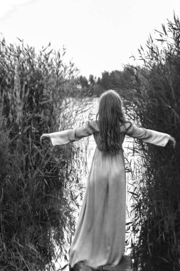 Sarışın, uzun elbiseli, gölün sularında ayakta duran, kamışların arasında elini kaldıran kadının arkası.