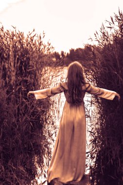 Sarışın, uzun elbiseli, gölün sularında ayakta duran, kamışların arasında elini kaldıran kadının arkası.