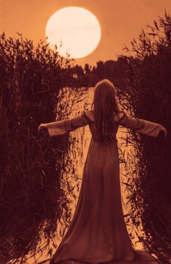 Sarışın, uzun elbiseli, gölün sularında ayakta duran, kamışların arasında elini kaldıran kadının arkası. Dolunay tepede..