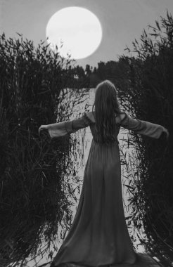 Sarışın, uzun elbiseli, gölün sularında ayakta duran, kamışların arasında elini kaldıran kadının arkası. Dolunay tepede..