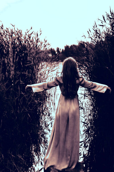 Back of blonde sensual woman in long dress standing in water of lake with raised hands between the reeds