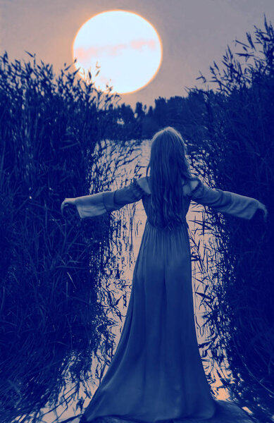 Back of blonde sensual woman in long dress standing in water of lake with raised hands between the reeds. full moon above head.