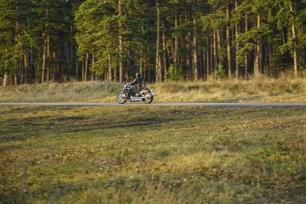 チョッパーバイクの若い男 美しい秋の森と緑の牧草地で空の道路に乗ってバイクに乗る — ストック写真