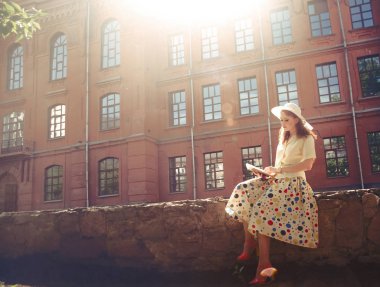 Eski çitlerin üzerinde oturan mutlu kızıl saçlı kız ve birçok penceresi olan kırmızı tuğla bina hakkında kitap okuyor. 