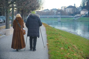 Yaşlı bir çift kışın parkta yürüyüşe çıkar; hem kadın hem de erkek alp yürüyüş direkleri kullanır. Soğuk kış gününde asfalt yolda Salzach (Salzburg, Avusturya) yakınlarında parkta yürüyen yaşlı çift. geri, arka plan.