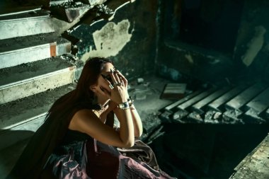 Yüzüne yakın el ele tutuşan soytarı kadın eski bir şatonun bodrumunun derinliklerinde karanlık bir taş merdivenin içinde oturuyor. Kabus, korku konsepti. Dairesel merdiven.