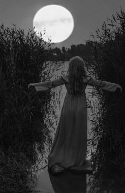 Sarışın, uzun elbiseli, gölün sularında ayakta duran, kamışların arasında elini kaldıran kadının arkası. Dolunay tepede..