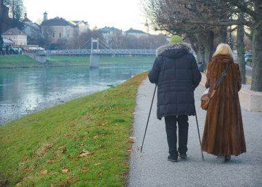 Yaşlı bir çift kışın parkta yürüyüşe çıkar; hem kadın hem de erkek alp yürüyüş direkleri kullanır. Soğuk kış gününde asfalt yolda Salzach (Salzburg, Avusturya) yakınlarında parkta yürüyen yaşlı çift. geri, arka plan.