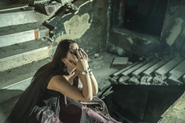 Yüzüne yakın el ele tutuşan soytarı kadın eski bir şatonun bodrumunun derinliklerinde karanlık bir taş merdivenin içinde oturuyor. Kabus, korku konsepti. Dairesel merdiven.