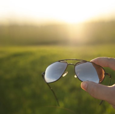 Erkek El, taze yeşil çim ya da çayır ve günbatımı arka planında güneş gözlüğü tutuyor. şık aksesuarlar. 