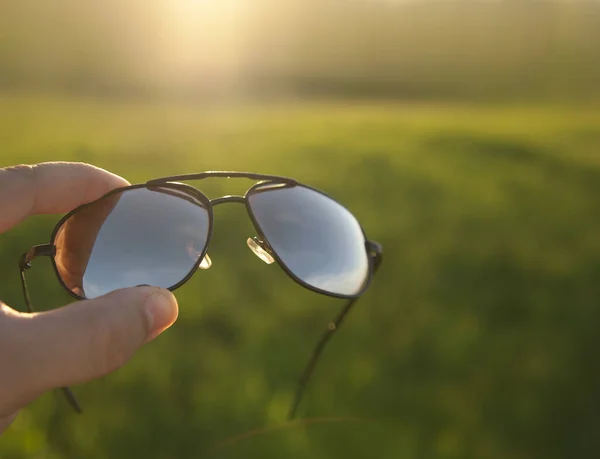 新鮮な緑の芝生のフィールドや牧草地や日没の空の背景に対する男性の手のサングラスを保持 スタイリッシュなアクセサリー — ストック写真