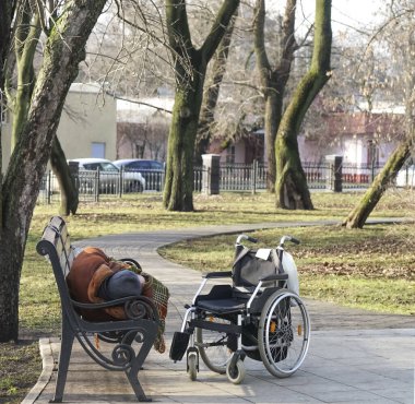 Soğuk baharda ya da sonbahar parkında bankta uzanmış dinlenen uyuyan son sınıf öğrencisi. Tekerlekli sandalye yakında duruyor. Adam kürk ve kalın şapka giyiyor..