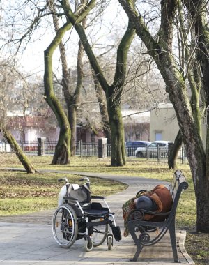 Soğuk baharda ya da sonbahar parkında bankta uzanmış dinlenen uyuyan son sınıf öğrencisi. Tekerlekli sandalye yakında duruyor. Adam kürk ve kalın şapka giyiyor..