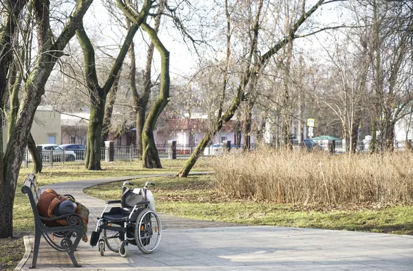 老年人在寒冷的春天或秋天的公园里躺在长椅上休息 站在旁边的轮椅 男人穿着毛皮外套 头戴暖和的帽子 — 图库照片