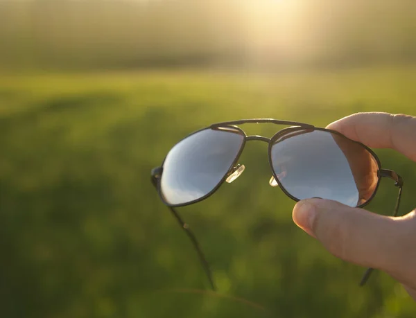 新鮮な緑の芝生のフィールドや牧草地や日没の空の背景に対する男性の手のサングラスを保持 スタイリッシュなアクセサリー — ストック写真