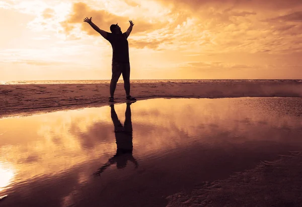 Silhouette of a young man with hands up against the background of the sunset in the reflection of the sea or pond. Silhouette of Man Raising His Hands or Open arms  with sunset, sunlight.