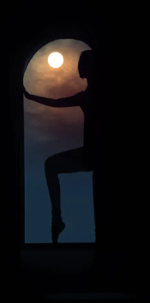 silhouette of a young good shaped woman standing on the huge window. Concept of full moon power. Beautiful girl standing between round window frame. midnight moon light.