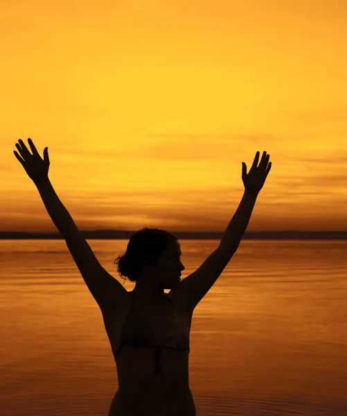 Woman silhouette on the beach at sunset. hands up. Girl standing hands up on the beach at sunset.Silhouette of a slender beautiful woman against the sunset on the sea. Sexy attractive figure of a woman standing on the beach in the rays of setting sun