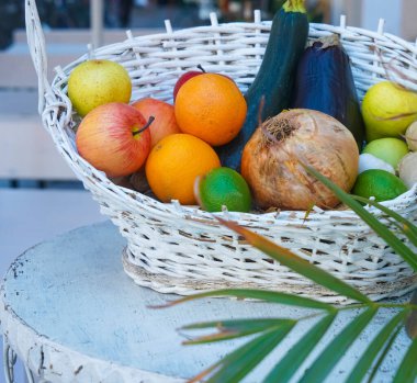 Tahta masa üzerinde limon, portakal ve elma bulunan çeşitli tropikal meyvelerden oluşan daha beyaz hasır sepet. Sepetteki sebzeler ve meyveler 