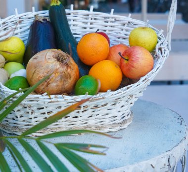 Tahta masa üzerinde limon, portakal ve elma bulunan çeşitli tropikal meyvelerden oluşan daha beyaz hasır sepet. Sepetteki sebzeler ve meyveler 