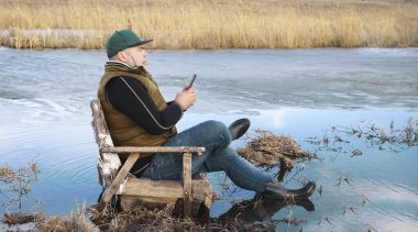 Olgun bir adamın bataklık kıyısındaki eski bir sandalyede cep telefonuyla dinlenmesinin yan görüntüsü. Göl rıhtımında oturan hippi adam cep telefonu kamerasıyla, rahat atmosferde, baharın başında fotoğraf çekiyor. lastik çizmeler