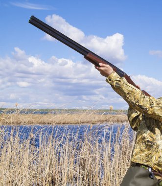Genç erkek av tüfeği ile çimenlerde oturuyor, avlandıktan sonra dinleniyor, palto ve lastik botlar giyiyor. Güneşli hava, sonbahar. Avcı adam bataklıkta kurban aramaya gider. Yan görünüm