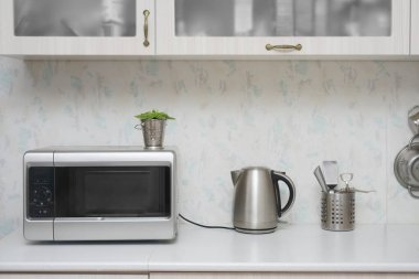 Yakın çekim mikrodalga fırın ve paslanmaz elektrikli su ısıtıcısı. Mutfakta beyaz, modern ahşap masa..