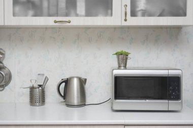 Yakın çekim mikrodalga fırın ve paslanmaz elektrikli su ısıtıcısı. Mutfakta beyaz, modern ahşap masa..