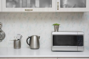 Yakın çekim mikrodalga fırın ve paslanmaz elektrikli su ısıtıcısı. Mutfakta beyaz, modern ahşap masa..