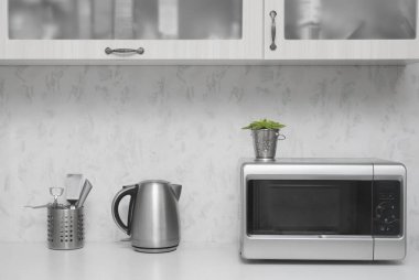 Yakın çekim mikrodalga fırın ve paslanmaz elektrikli su ısıtıcısı. Mutfakta beyaz, modern ahşap masa..