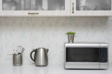 Yakın çekim mikrodalga fırın ve paslanmaz elektrikli su ısıtıcısı. Mutfakta beyaz, modern ahşap masa..