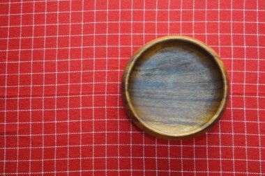 Beyaz tahta masanın üzerinde kırmızı gingham masa örtüsü olan boş yuvarlak ahşap bir tabak. En üst görüntü.