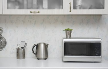 Yakın çekim mikrodalga fırın ve paslanmaz elektrikli su ısıtıcısı. Mutfakta beyaz, modern ahşap masa..