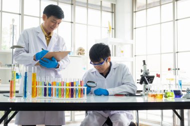 Bilim, Kimya, Teknoloji, Biyoloji ve Laboratuar Konsepti - Asyalı kıdemli bilim adamı notlar alırken, Asyalı kıdemsiz bilim adamı petri kabıyla ilgili bir şeye bakıyor.