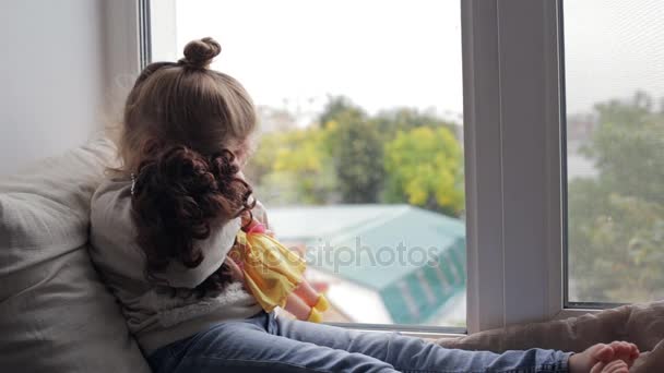 Menina bonita com boneca sentada em um peitoril da janela — Vídeo de Stock