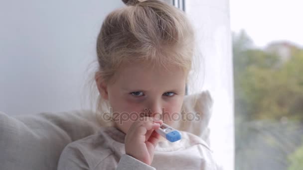 Ung sjuk flicka mäter temperaturen nära fönstret. Holding termometer i munnen — Stockvideo