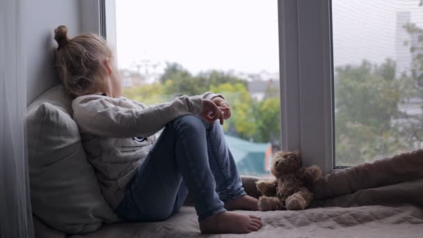 Sad cute young girl sitting on a window sill — Stock Video