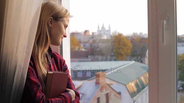 Piękna młoda kobieta z książki patrząc przez okno — Wideo stockowe
